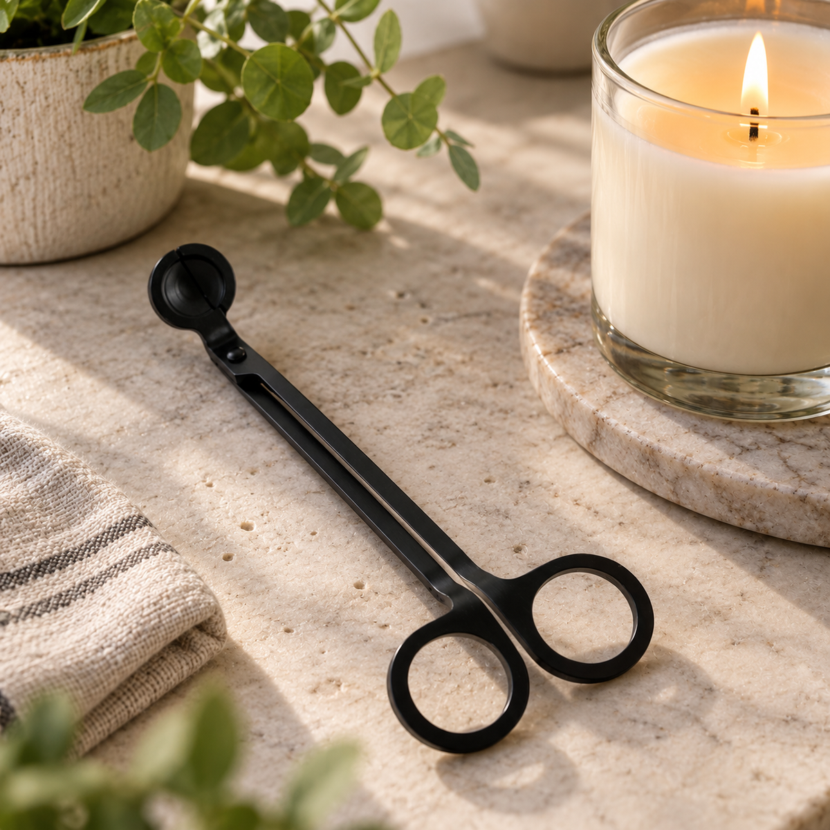 Matte black candle wick trimmer lying on a stone countertop with greenery and a candle in the background