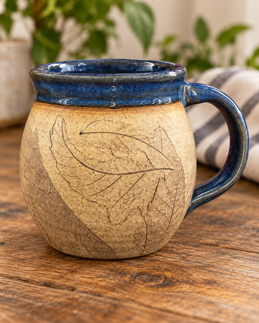 Front of a handmade Julia E. Dean stoneware mug with pressed leaf prints and a glossy deep-blue rim and handle