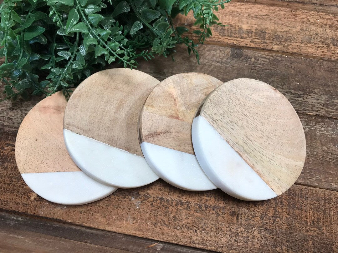 Set of four round mango wood and white marble coasters on a wooden table with a plant in the background