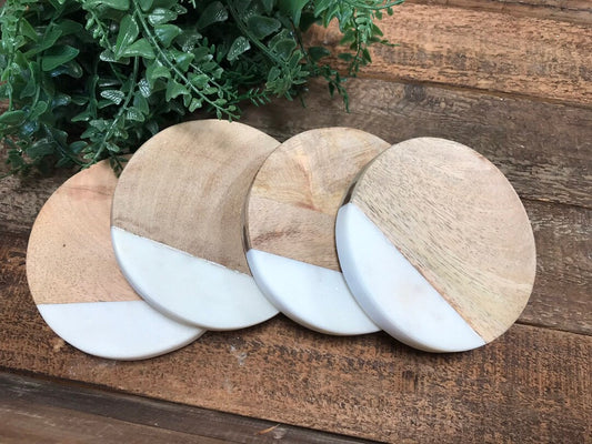 Set of four round mango wood and white marble coasters on a wooden table with a plant in the background