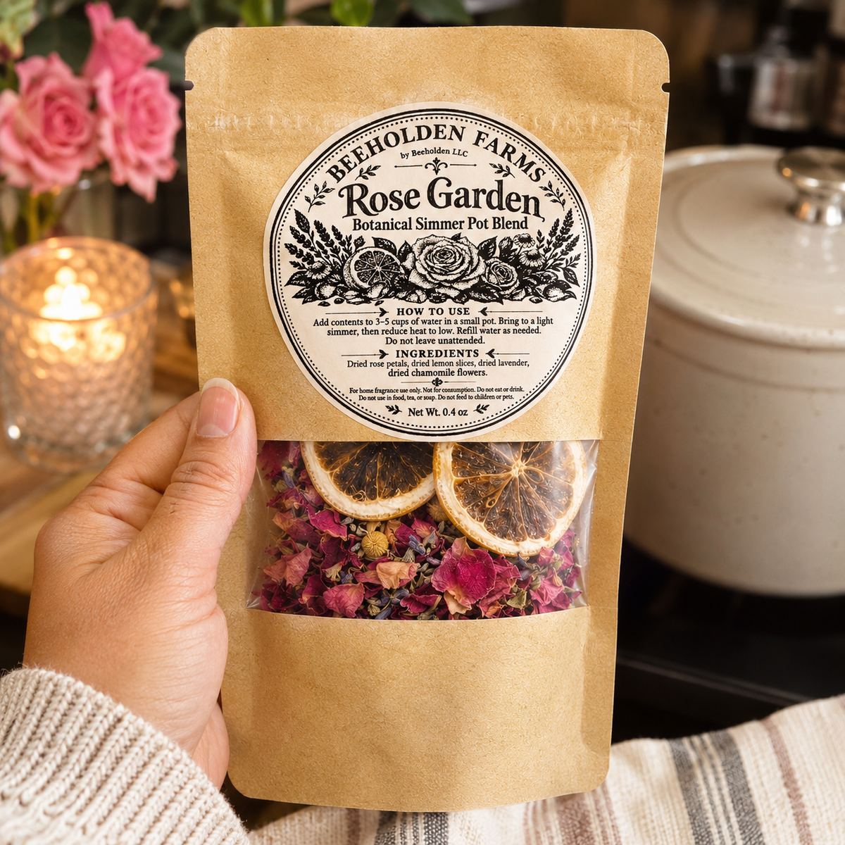 Hand holding a Rose Garden botanical simmer pot blend pouch by Beeholden Farms in a cozy kitchen with fresh roses and a candle